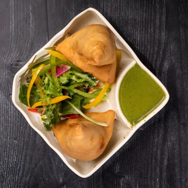 Two golden-brown samosas with a fresh green salad and vibrant green chutney on a white plate.