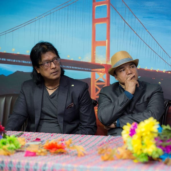 Two men in suits sit at a table with colorful decorations, a Golden Gate Bridge backdrop behind them.